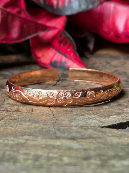 Copper bracelet with floral patterns on a wooden surface with red leaves in the background 