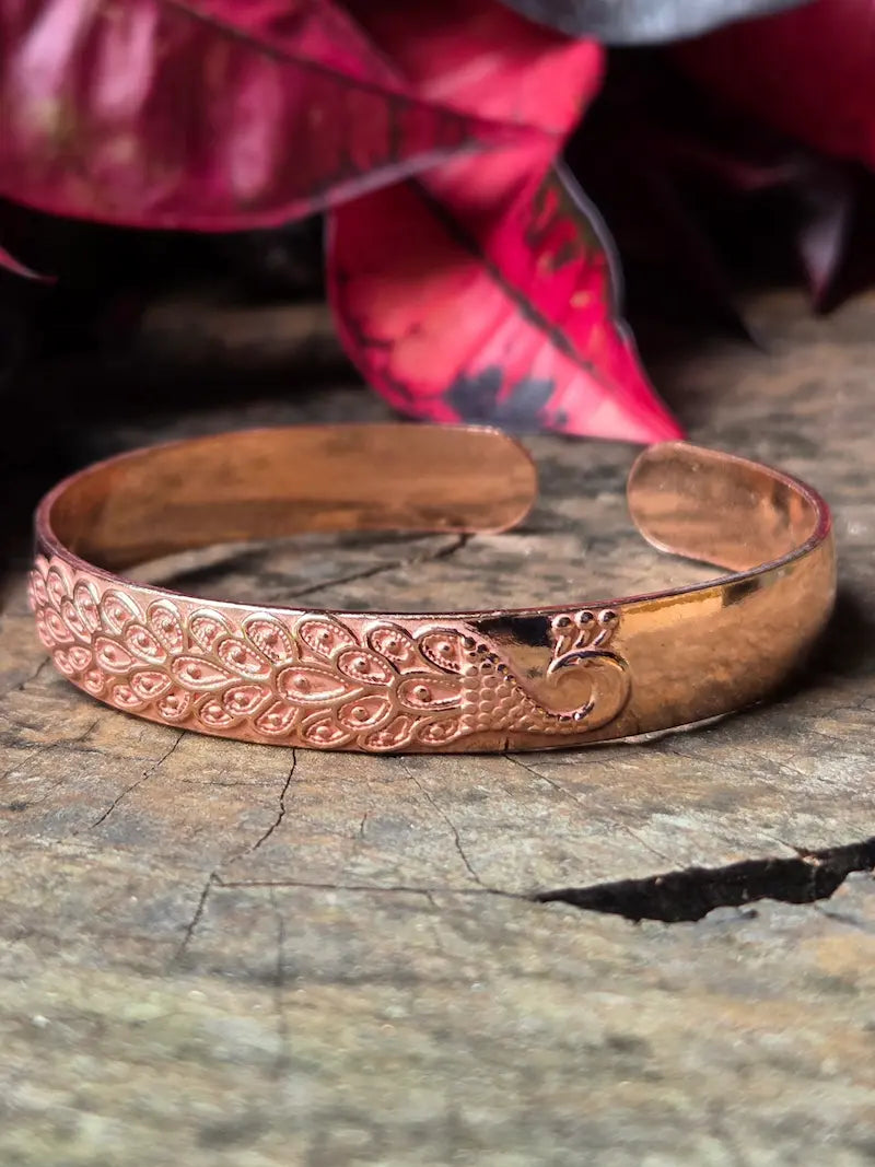 Copper bracelet with a peacock design on a wooden surface with red leaves in the background