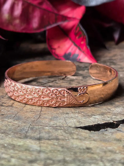 Copper bracelet with a peacock design on a wooden surface with red leaves in the background