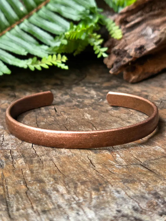 A copper cuff bracelet resting on a rustic wood surface with green fern leaves and a piece of driftwood in the background.