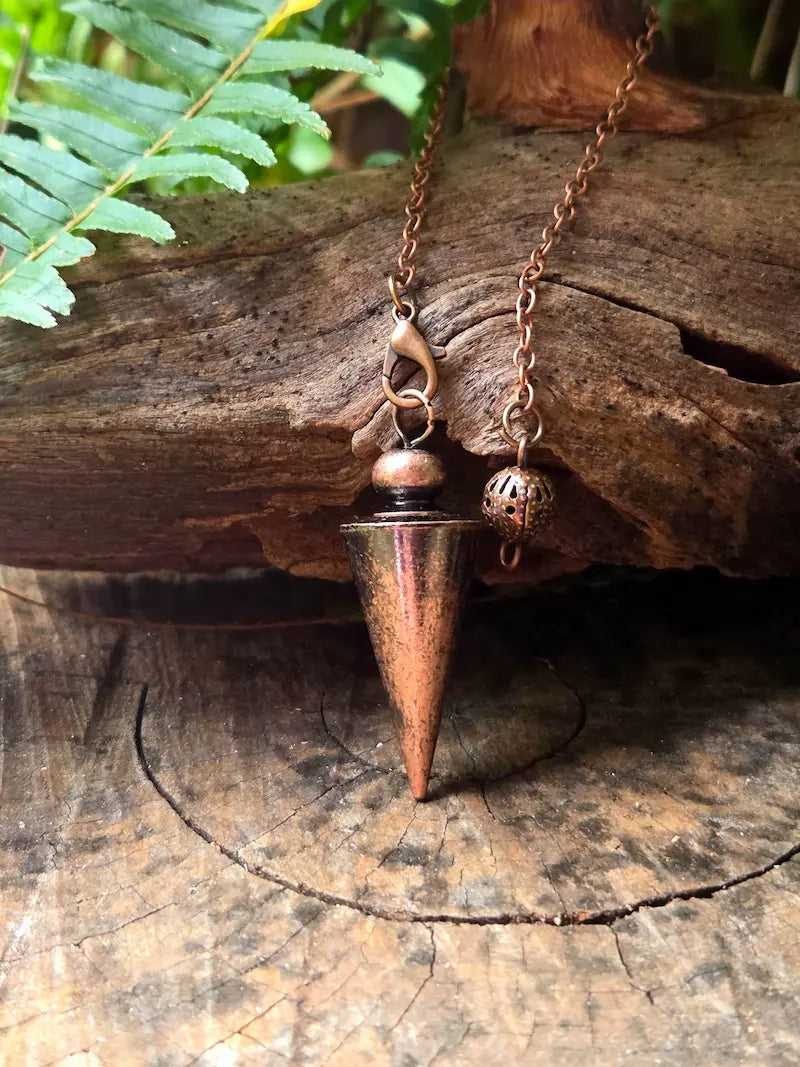Copper pendulum on a natural wooden surface with green ferns in the background