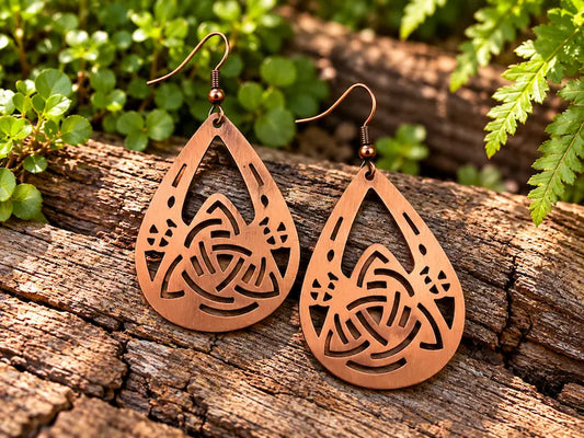Copper Earrings with a celtic knot design, resting on a natural wooden surface with green leaves in the background 