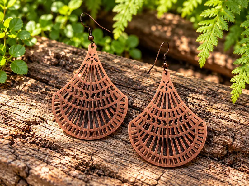 Copper Earrings with a filigree fan design, resting on a natural wooden surface with green leaves in the background 