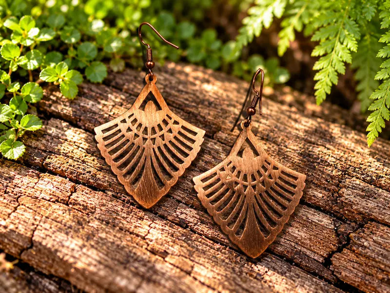 Copper Earrings with a geometric fan design, resting on a natural wooden surface with green leaves in the background 