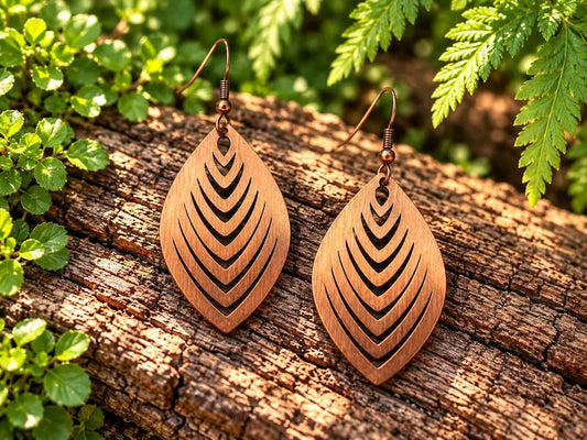 Copper earrings with a leaf design, resting on a natural wooden surface with green leaves in the background 