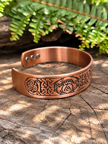 A copper-toned engraved cuff bracelet with a celtic knot design, displayed on a rustic wood surface with green fern leaves in the background.