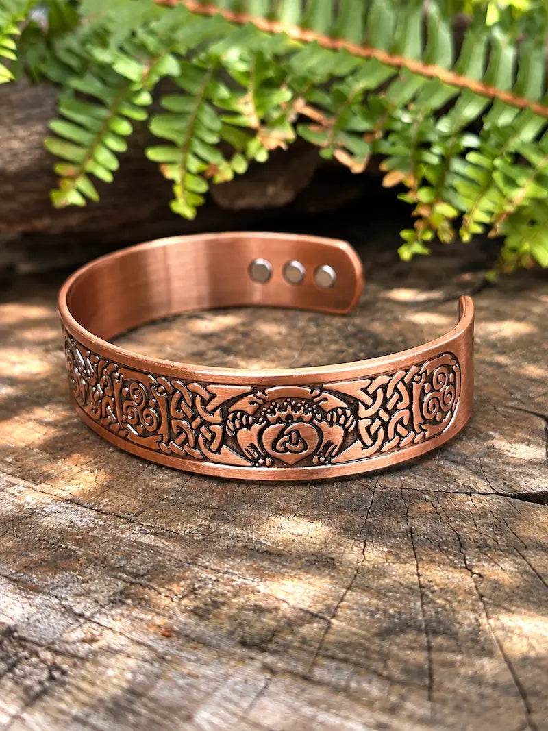 A copper-toned engraved cuff bracelet with a celtic knot design, displayed on a rustic wood surface with green fern leaves in the background.