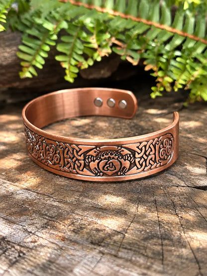 A copper-toned engraved cuff bracelet with a celtic knot design, displayed on a rustic wood surface with green fern leaves in the background.