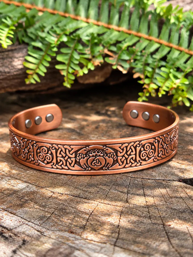 A copper-toned engraved cuff bracelet with a celtic knot design, displayed on a rustic wood surface with green fern leaves in the background.