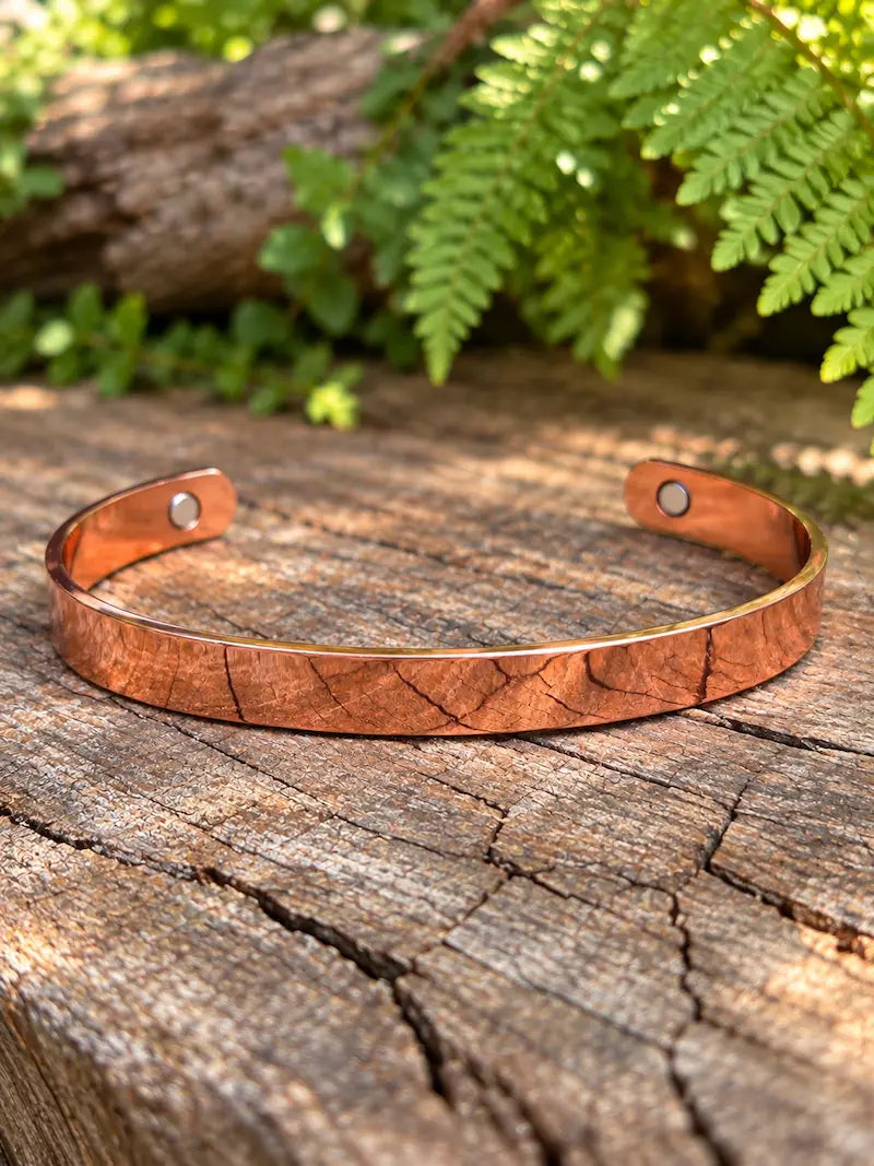 A copper cuff bracelet with a polish finish, resting on a weathered wooden log with green ferns in the background.