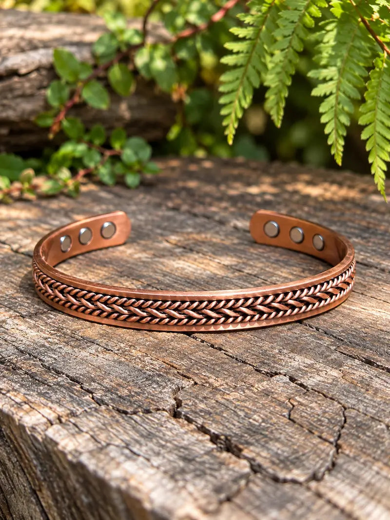 A copper bracelet with a twisted design, resting on a natural wooden surface with green ferns in the background 