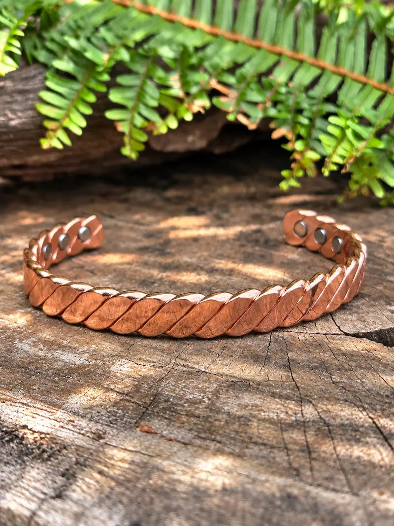 A polished copper cuff bracelet with a twisted copper design, displayed on a rustic wooden surface with green fern leaves in the background.