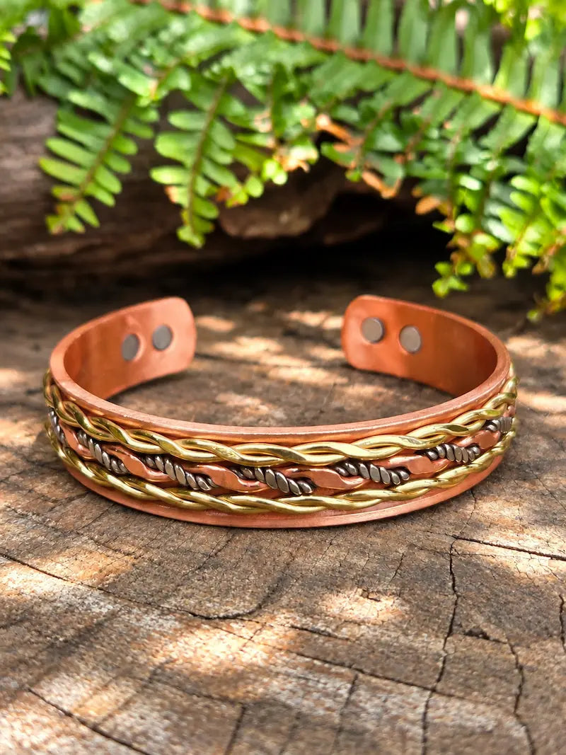 A handmade copper cuff bracelet with braided gold, silver and copper wire detailing, displayed on a rustic wooden log with green ferns in the background.