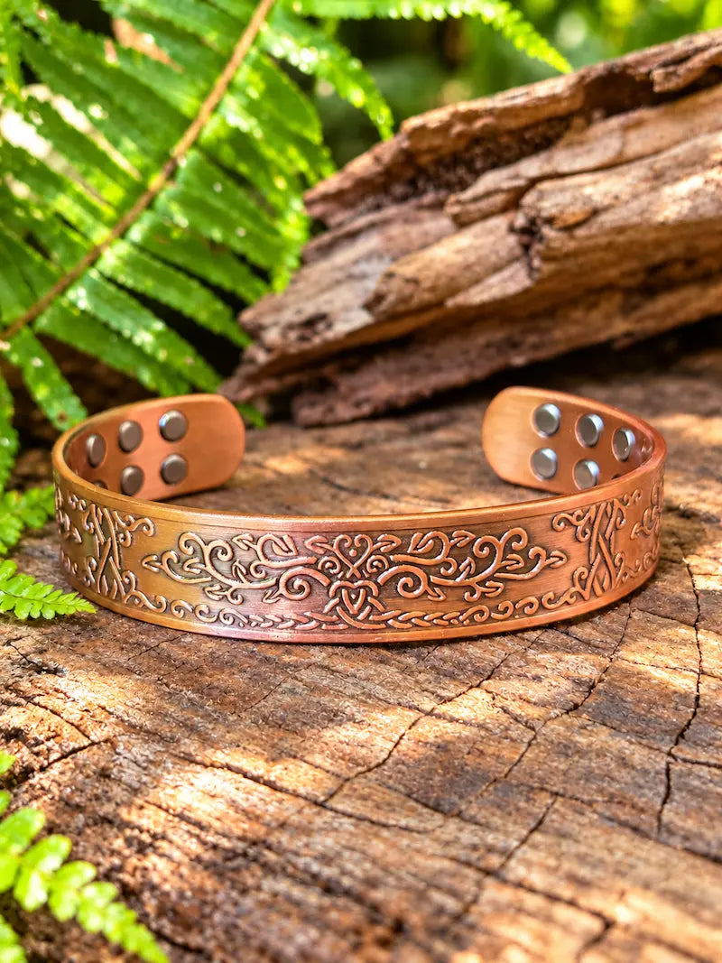 Copper bracelet with a vine scrollwork design, resting on a natural wooden surface with green ferns the background 