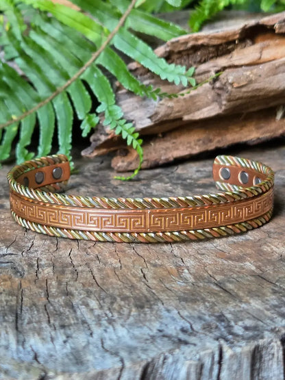 A copper bracelet with a Tricolour greek key design,  resting on a natural wooden surface with green ferns in the background.