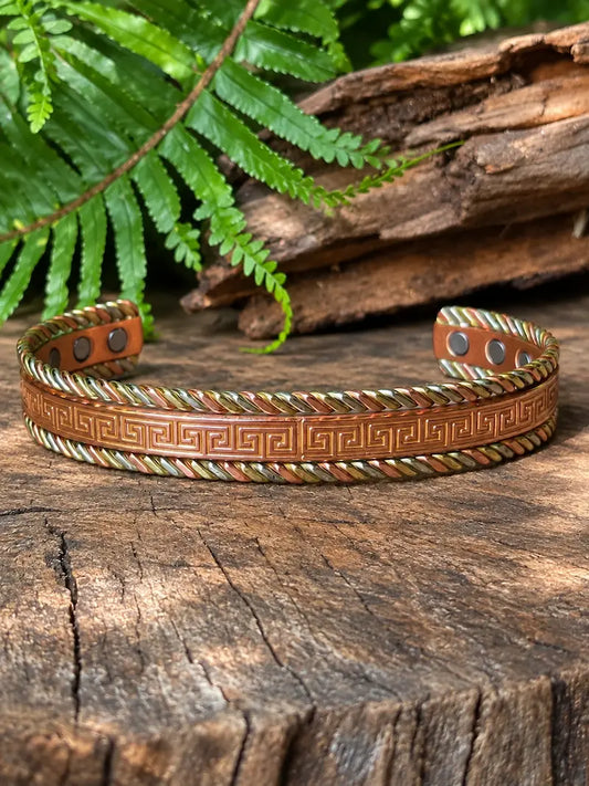 A copper bracelet with a Tricolour greek key design,  resting on a natural wooden surface with green ferns in the background.