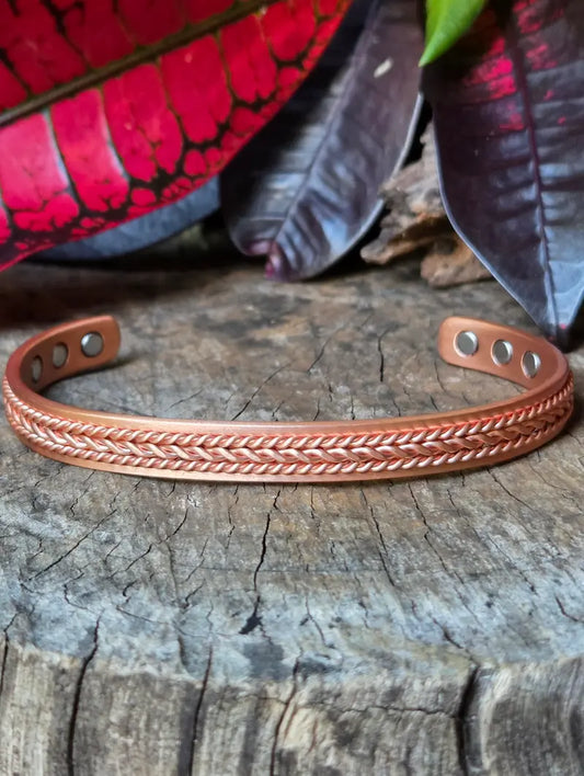 A copper bracelet with a twisted design, resting on a natural wooden surface with red leaves in the background.