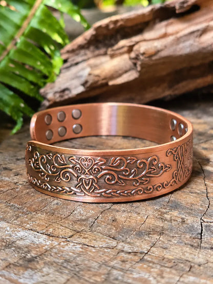 Copper bracelet with a vine scrollwork design, resting on a natural wooden surface with green ferns the background 