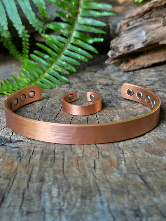A plain copper bracelet and ring resting on a natural wooden surface with green ferns in the background 