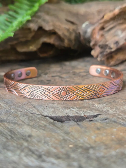 Copper bracelet with Geometric designs on a natural wooden surface with green ferns in the background 