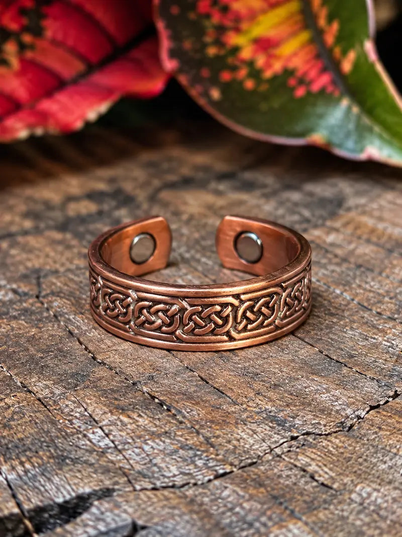 A copper ring with a celtic knot design, resting on a natural wooden surface with red leaves in the background 