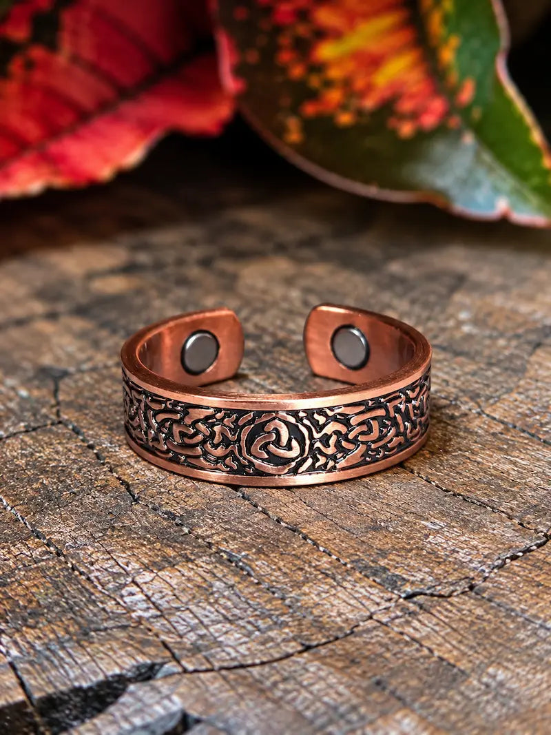 A copper ring with a celtic knot design, resting on a natural wooden surface with red leaves in the background 