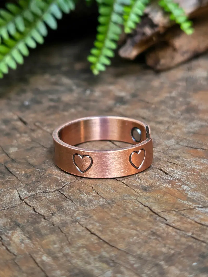 Copper ring with a love heart design on a natural wooden surface with green leaves in the background 