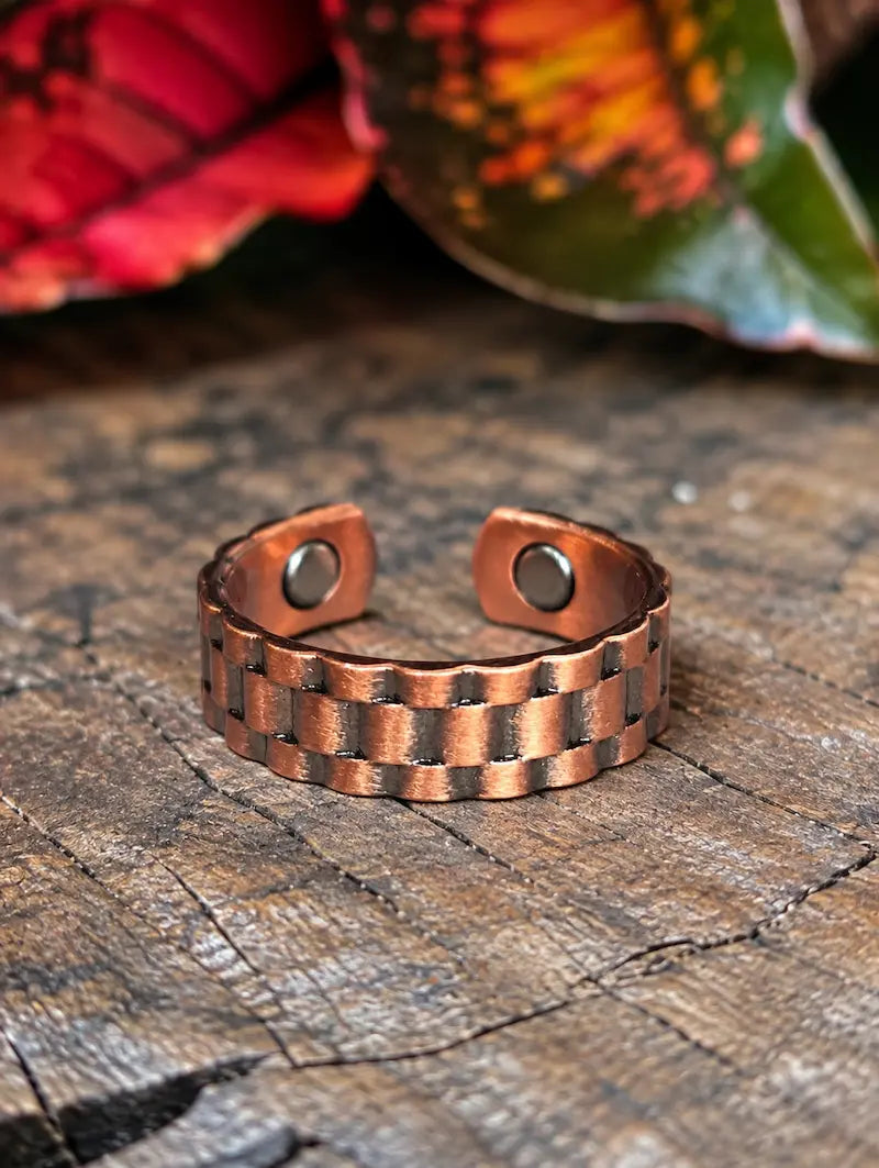 A copper ring with a link design, resting on a natural wooden surface with red leaves in the background 