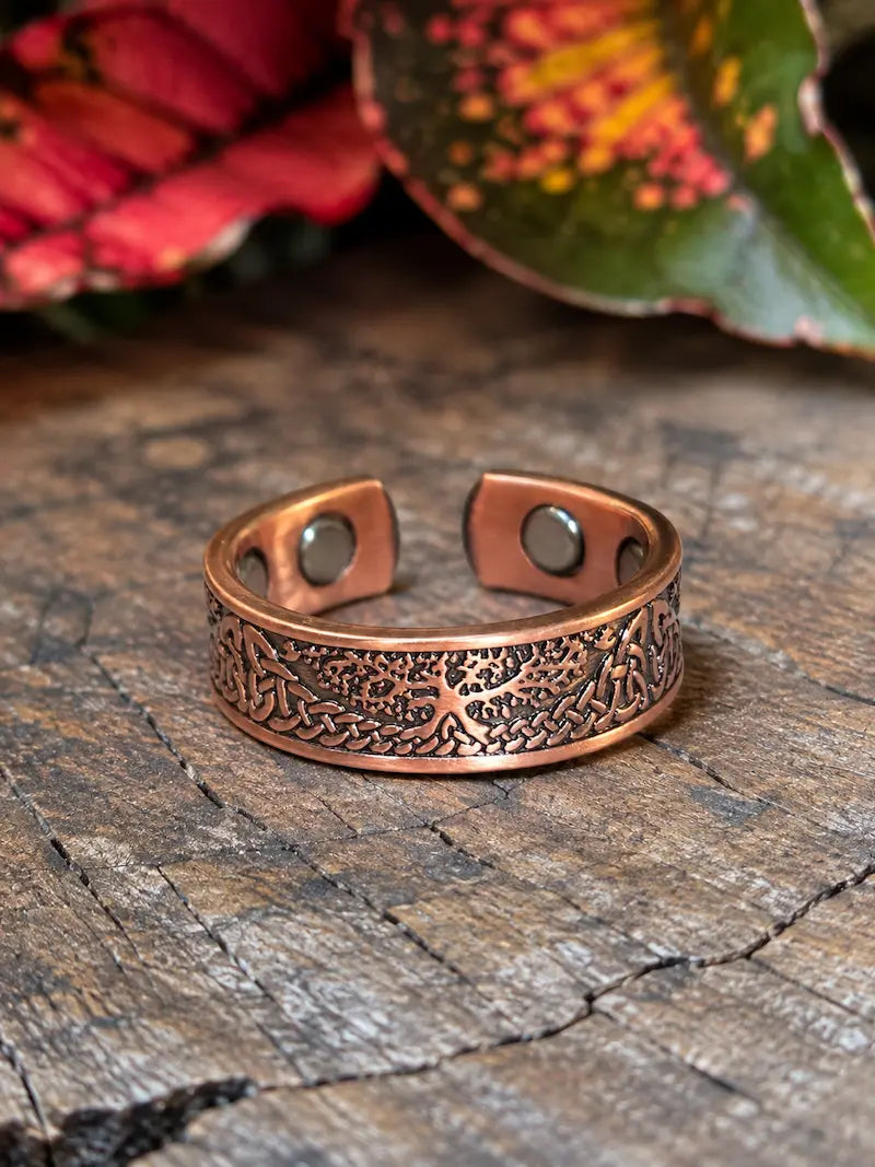 A copper ring with a tree of life design, resting on a natural wooden surface with red leaves in the background 