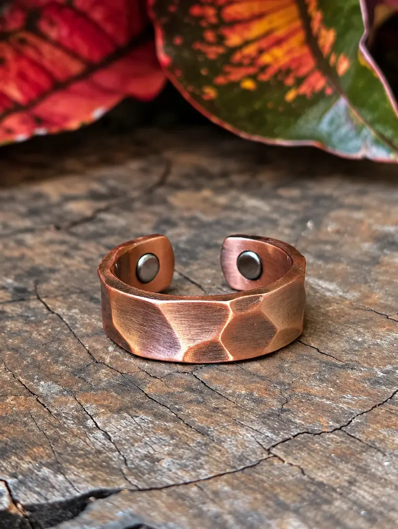 A hammered copper ring with two magnets, resting on a textured wooden surface with blurred red and green leaves in the background.