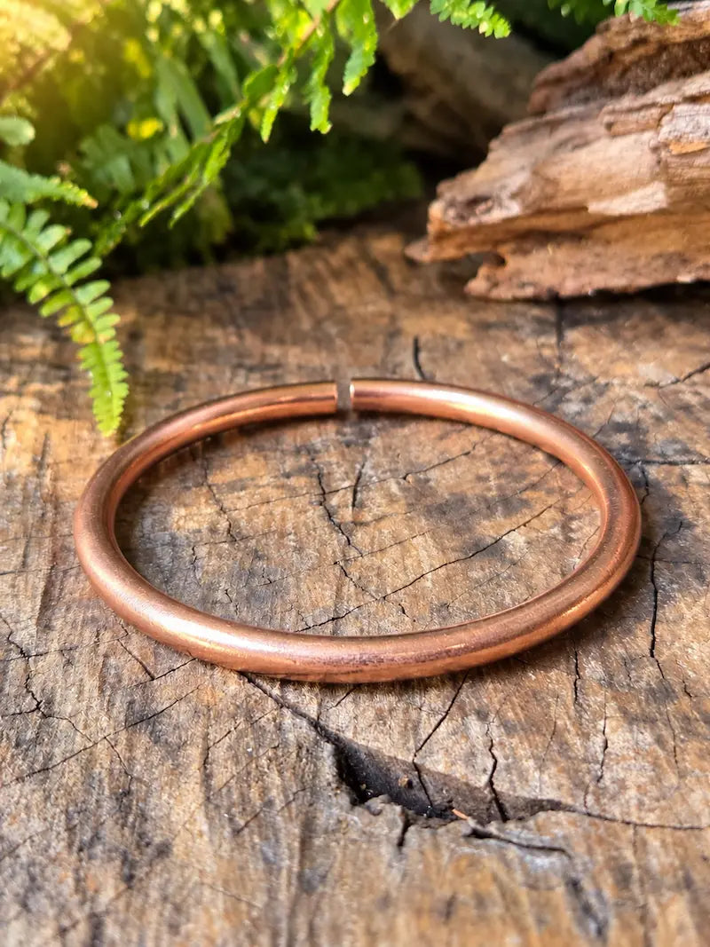 Copper bracelet with a open cuff bangle design, resting on a natural wooden surface with green ferns in the background 