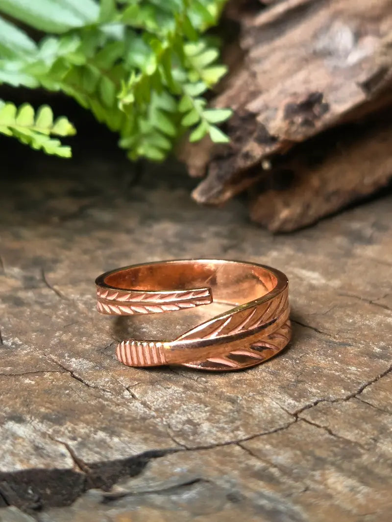 A copper ring with textured leaf-like detailing, resting on a rustic wood surface with blurred green leaves and bark in the background.