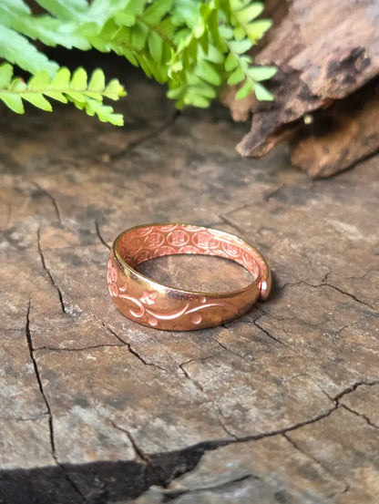 A pure copper ring with engraved floral patterns, resting on a textured wooden surface with green fern leaves in the background.