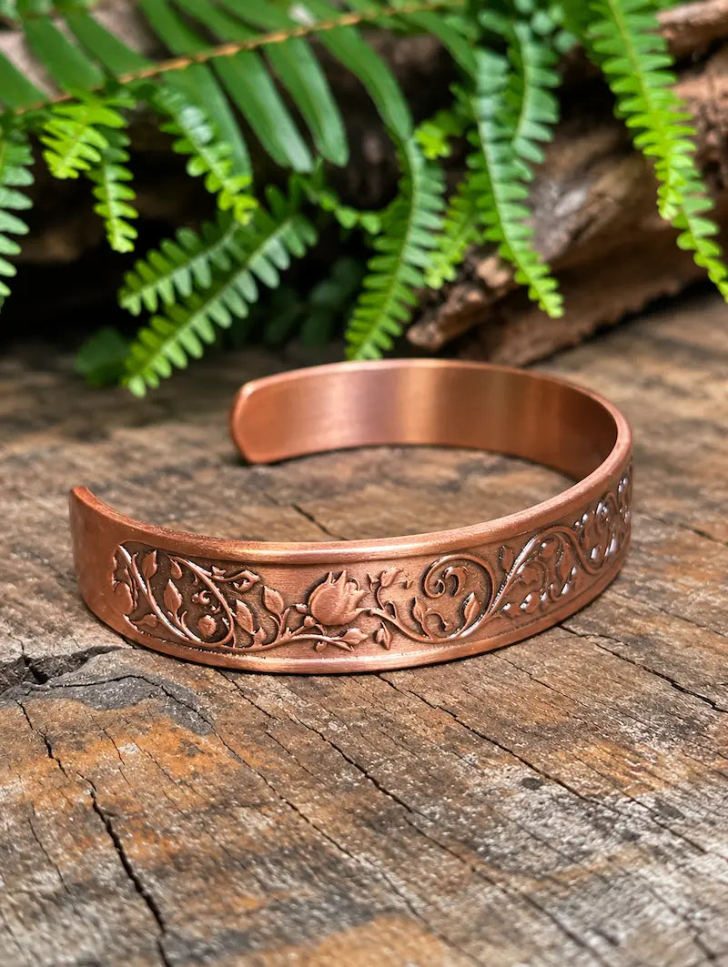 A copper cuff bracelet with a floral rose design, resting on a natural wooden surface with green leaves in the background 
