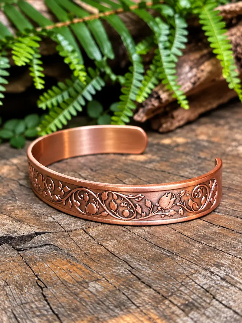 A copper cuff bracelet with a floral rose design, resting on a natural wooden surface with green leaves in the background 