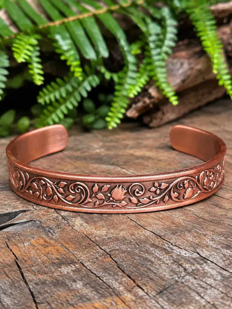 A copper cuff bracelet with a floral rose design, resting on a natural wooden surface with green leaves in the background 