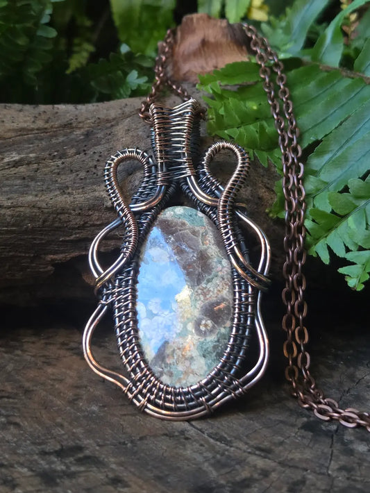 Copper wire-wrapped pendant with a natural stone on a wooden surface with greenery.