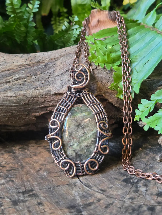 Copper necklace with a green stone pendant on a wooden surface with ferns in the background
