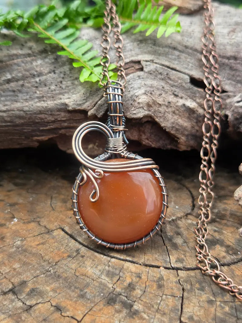 Copper wire wrapped pendant necklace with a red agate stone, resting on a natural wooden surface with green ferns in the background 