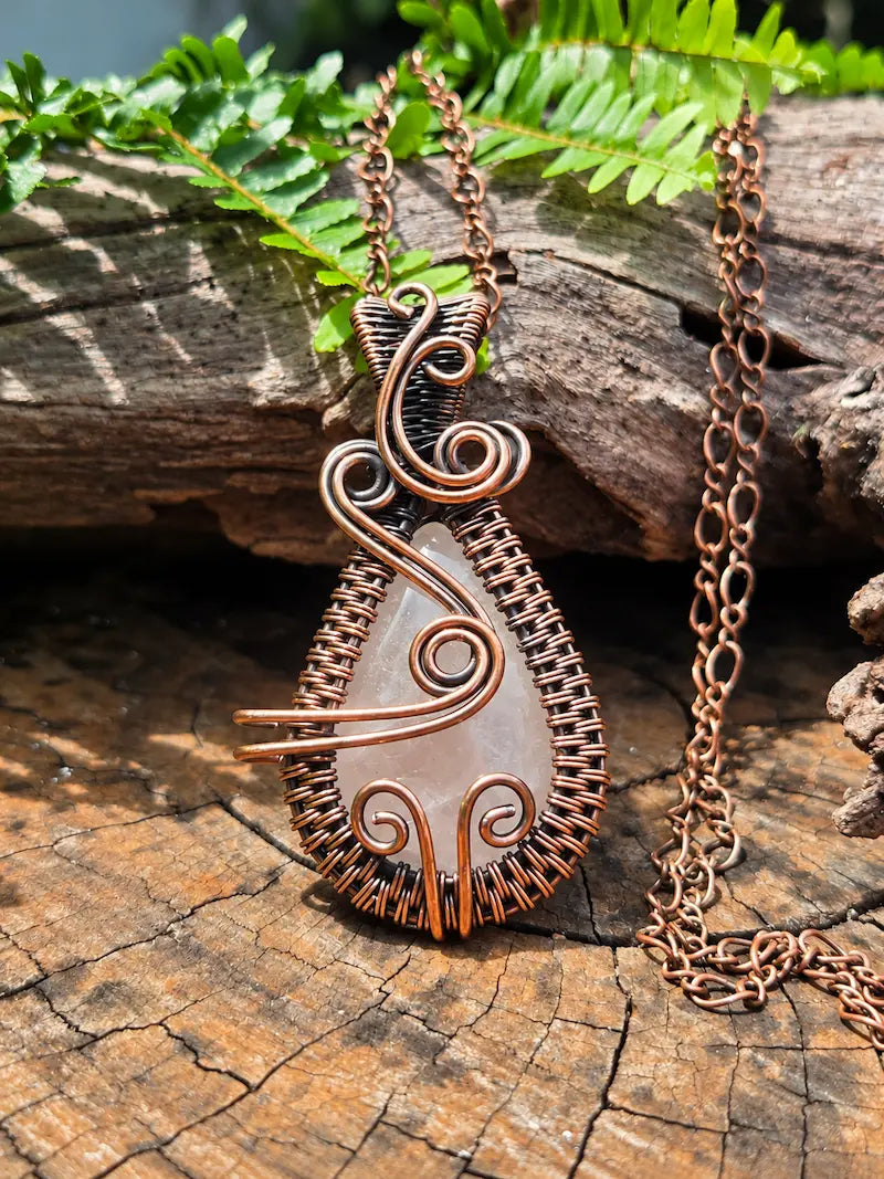 Copper pendant with a rose quartz crystal, resting on a natural wooden surface with green ferns in the background 