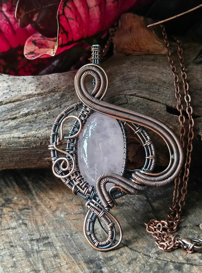 A handmade copper wire-wrapped pendant necklace featuring a polished pale pink stone, set in a rustic environment on a piece of weathered wood with red leaves.