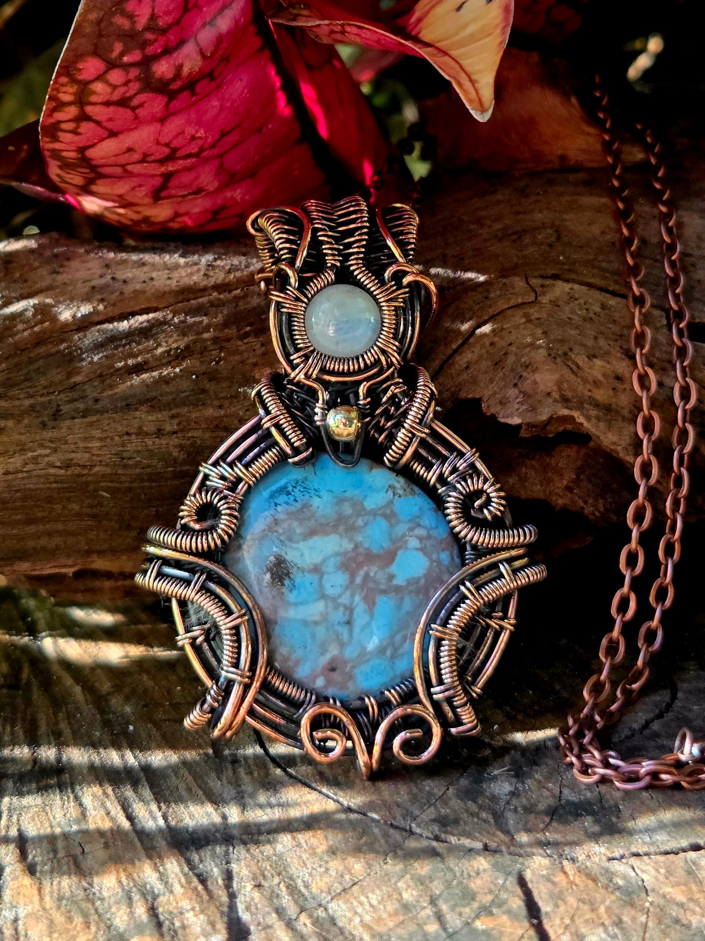 Turquoise and moonstone copper pendant necklace on a natural wooden surface with red leaves in the background 