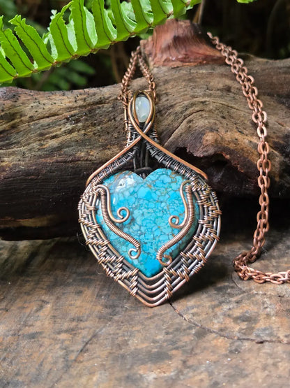 Turquoise heart-shaped pendant with copper wirework on a wooden surface with fern leaves.