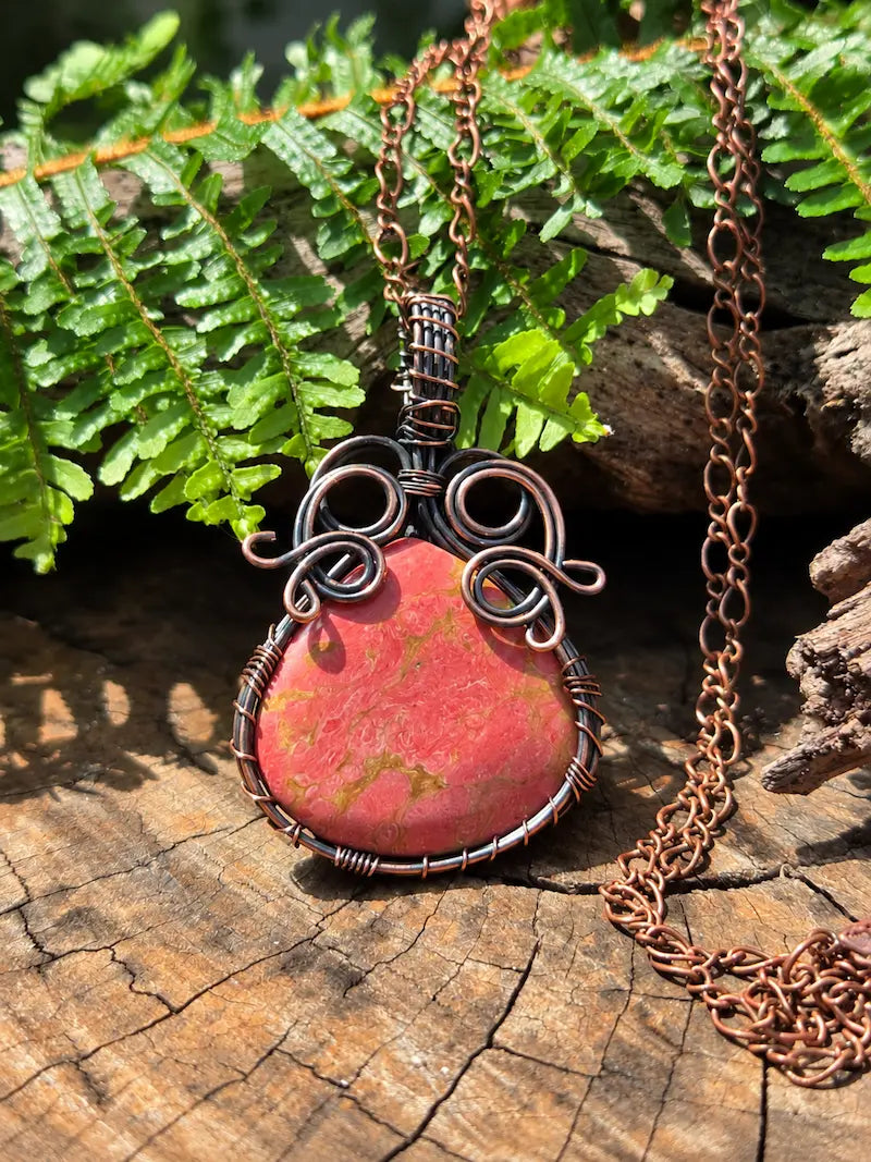 Copper pendant with a unakite stone, resting on a natural wooden surface with green ferns in the background 