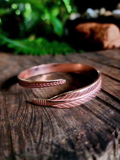 A copper feather bracelet placed on a wooden surface with blurred greenery in the background.