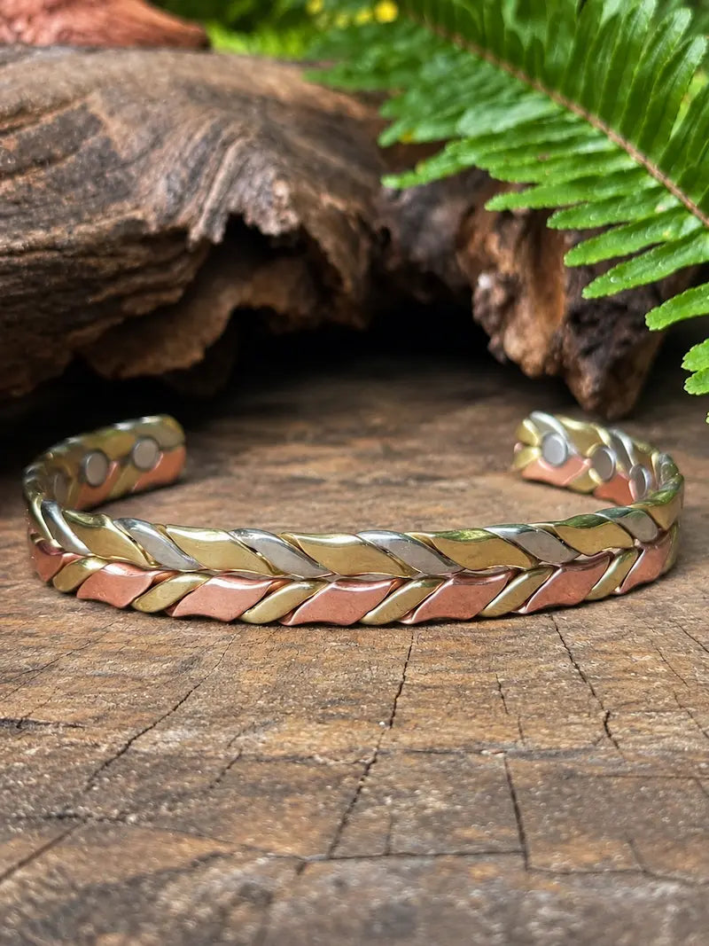 A braided cuff bracelet with alternating metallic rose gold, gold, and silver bands, displayed on a rustic wooden surface with a fern leaf in the background.