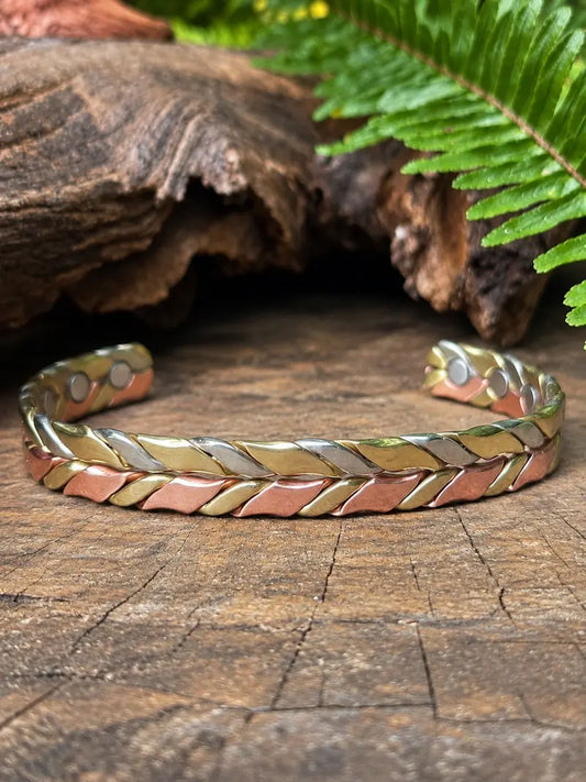 A braided cuff bracelet with alternating metallic rose gold, gold, and silver bands, displayed on a rustic wooden surface with a fern leaf in the background.