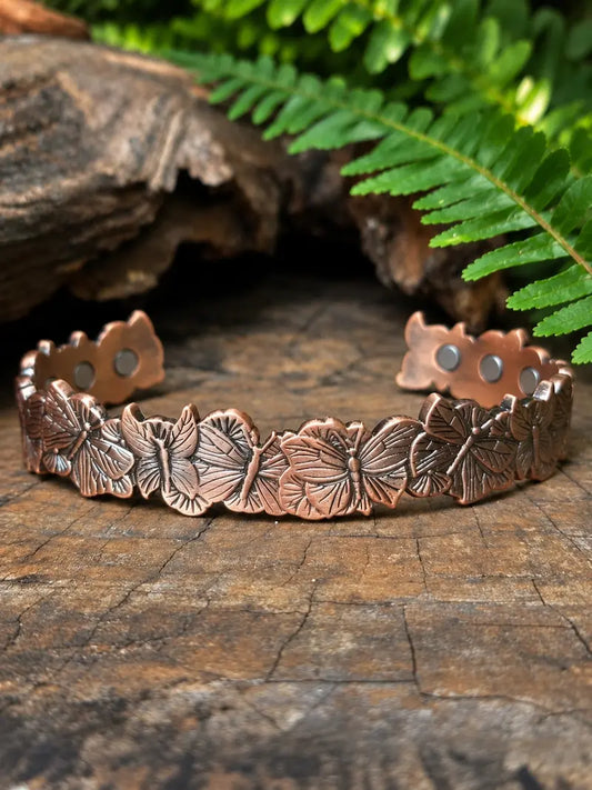 Copper bracelet with butterfly patterns on a wooden surface with ferns in the background