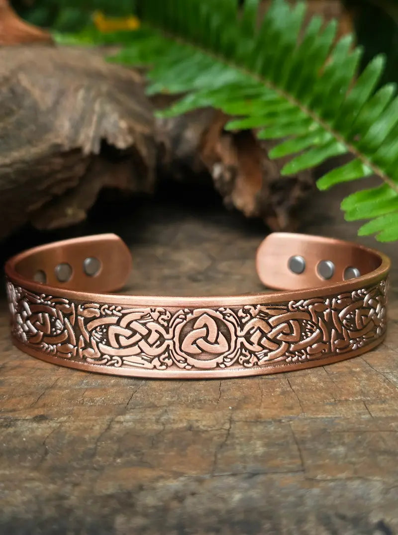 Copper bracelet with intricate designs on a wooden surface with ferns in the background