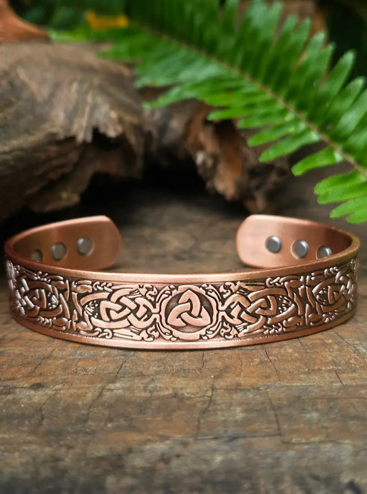 Copper bracelet with intricate designs on a wooden surface with ferns in the background